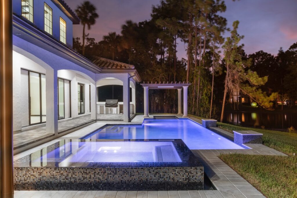 Modern backyard with illuminated swimming pool, hot tub, covered patio, and pergola next to a two-story house at dusk, surrounded by trees.