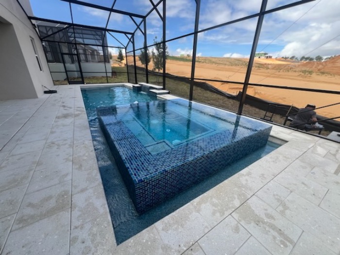 A modern backyard pool with a built-in, raised spa surrounded by blue mosaic tiles, enclosed by a screened structure with views of a sandy landscape outside.