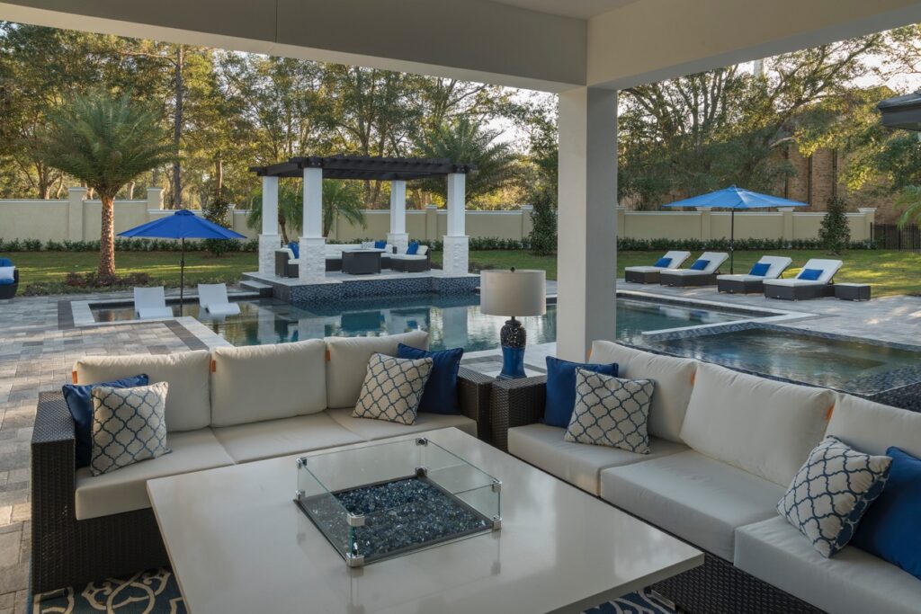 Modern outdoor patio with white sectional sofas, blue accent pillows, a fire pit table, and a pool area with loungers, umbrellas, and a covered seating area in the background.