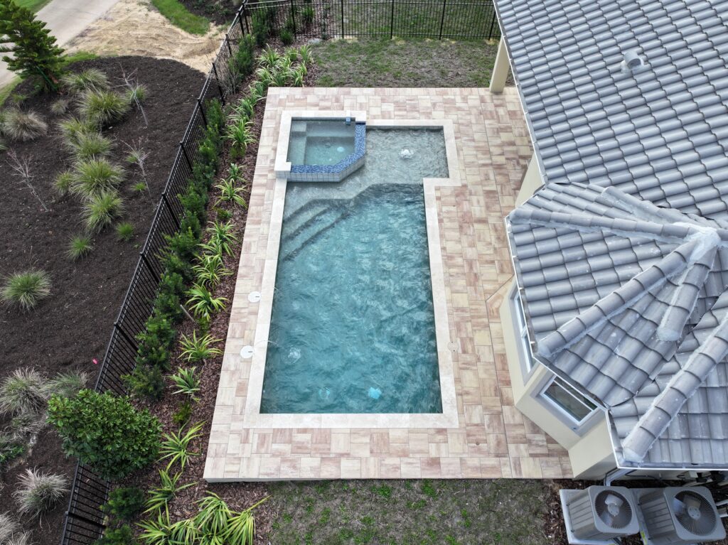 A rectangular backyard swimming pool with an attached hot tub, bordered by tan tiles, next to a house with a gray tiled roof and surrounded by landscaping.