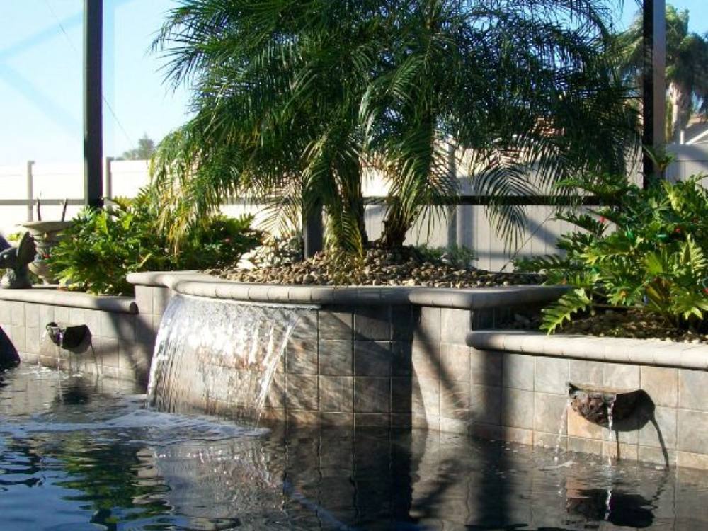 A small tiled waterfall feature spills into a pool, with a palm tree and green plants growing in a raised planter behind it.