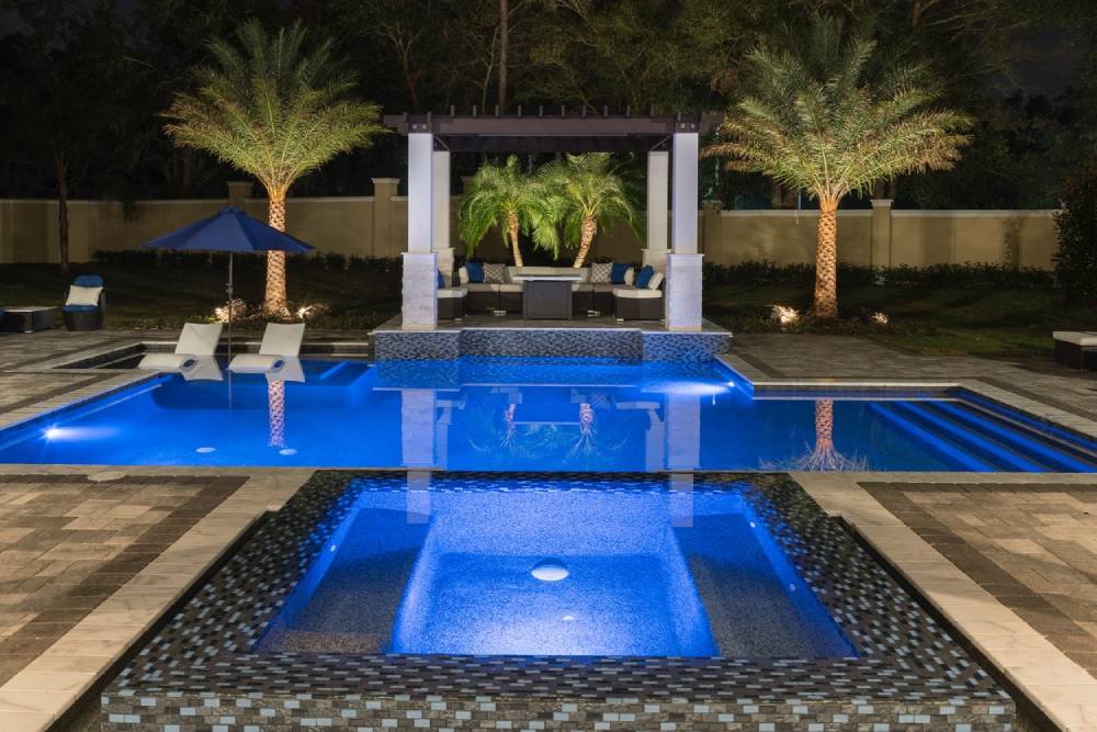 Modern backyard pool at night with illuminated water, hot tub in the foreground, lounge chairs in the water, a pergola, palm trees, and patio seating in the background.