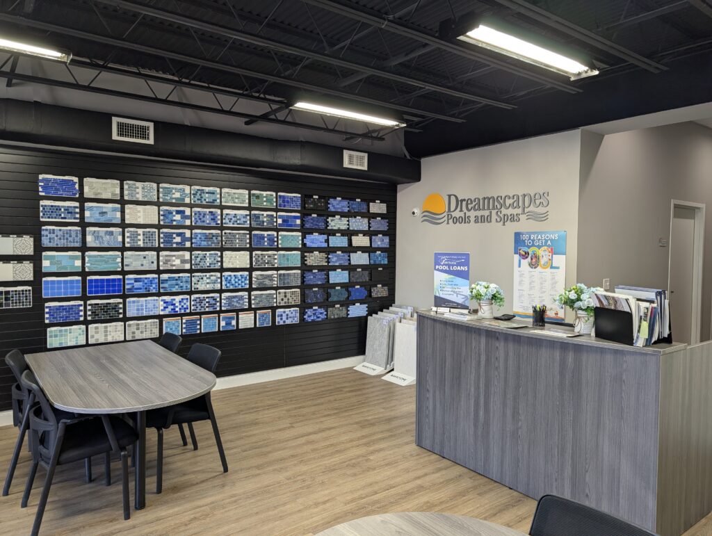 Interior of a pool and spa showroom with a wall display of tile samples, a reception desk, tables with chairs, and promotional materials on the countertop.