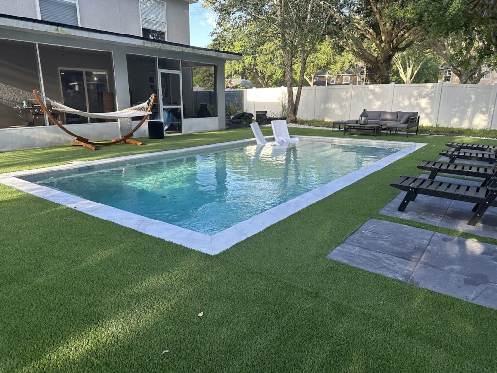 Backyard with a rectangular swimming pool, two in-pool lounge chairs, artificial grass, lounge chairs, a hammock, and patio furniture near a white house.