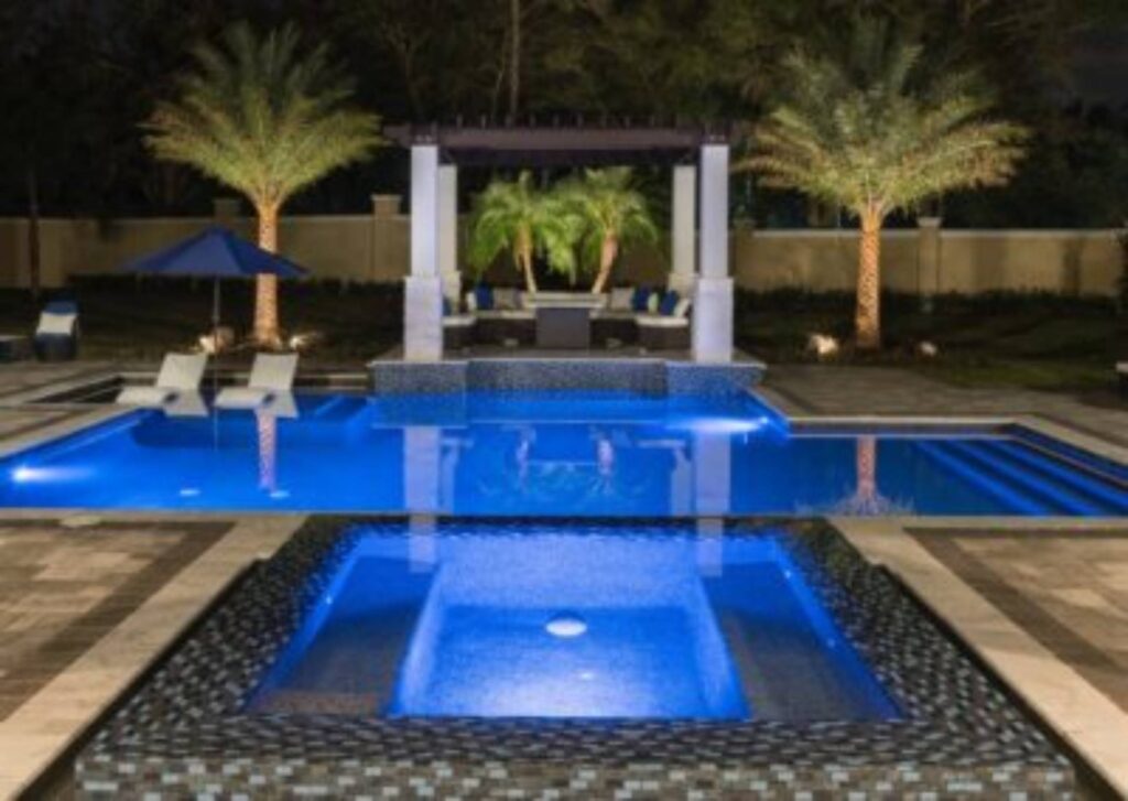 A modern backyard pool area at night with illuminated blue water, a hot tub in the foreground, lounge chairs, palm trees, and a pergola with seating in the background.