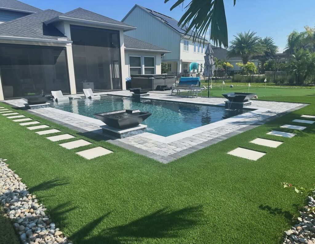 Modern backyard with a rectangular pool expertly crafted by Pool Builders in Lake Nona, lounge chairs in the water, stepping stones on artificial grass, and contemporary landscaping under a sunny sky.