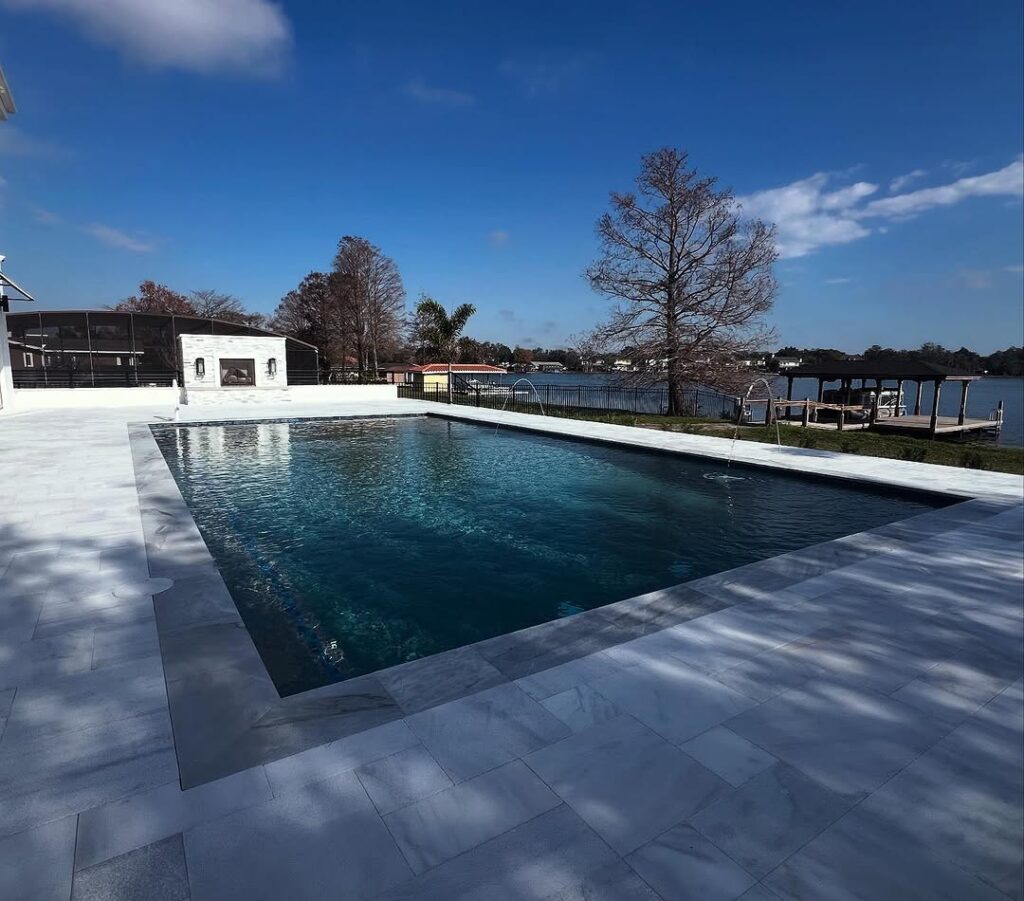 A rectangular outdoor swimming pool, expertly crafted by Pool Builders in Clermont, features light-colored tile surrounds and overlooks a lake with a dock and trees in the background under a partly cloudy sky.