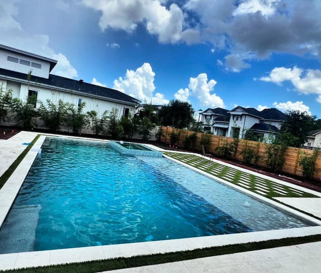 Rectangular outdoor swimming pool with clear blue water, crafted by expert Pool Builders in Lake Nona, surrounded by stone decking, grass strips, and a wooden fence; houses and clouds visible in the background.