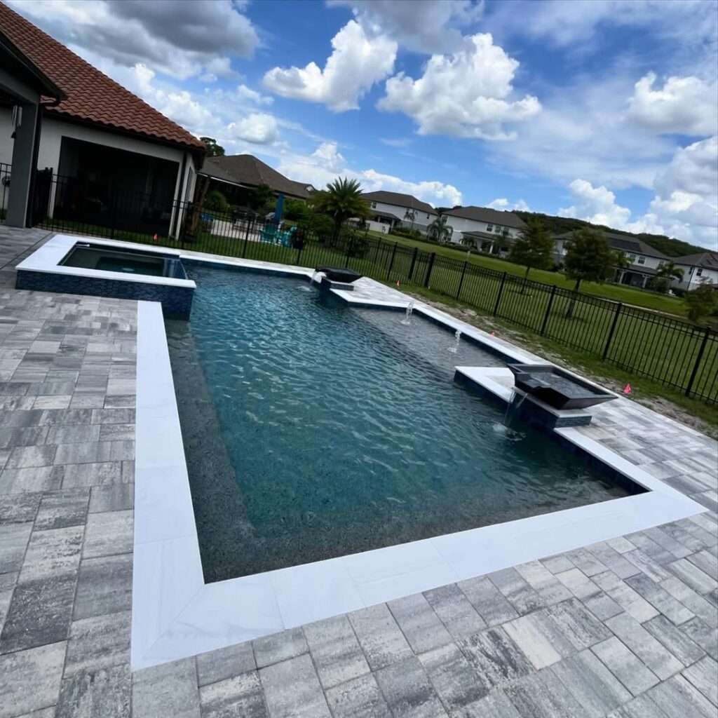 A rectangular backyard swimming pool with a hot tub, expertly crafted by Pool Builders in Lake Nona, is surrounded by light gray stone tiles near a fenced lawn and neighboring houses under a partly cloudy sky.