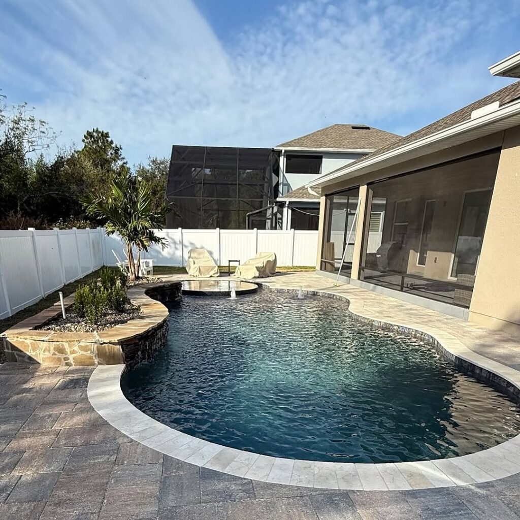 Backyard with a curved swimming pool designed by Pool Builders in Clermont, stone patio, lounge chairs, small palm tree, and a screened porch attached to the house.