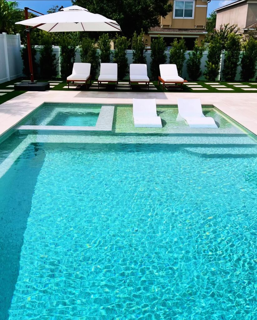 A backyard pool by Pool Builders in Lake Nona features two white lounge chairs in the water, four poolside loungers under an umbrella, and a hot tub area, all surrounded by lush greenery and a house in the background.