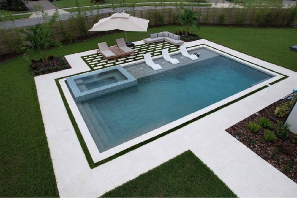A modern backyard by skilled Pool Contractors in Orlando features a rectangular pool with built-in loungers, a connected hot tub, a patio area with a sectional sofa and umbrella, all surrounded by lush lawn and landscaping.