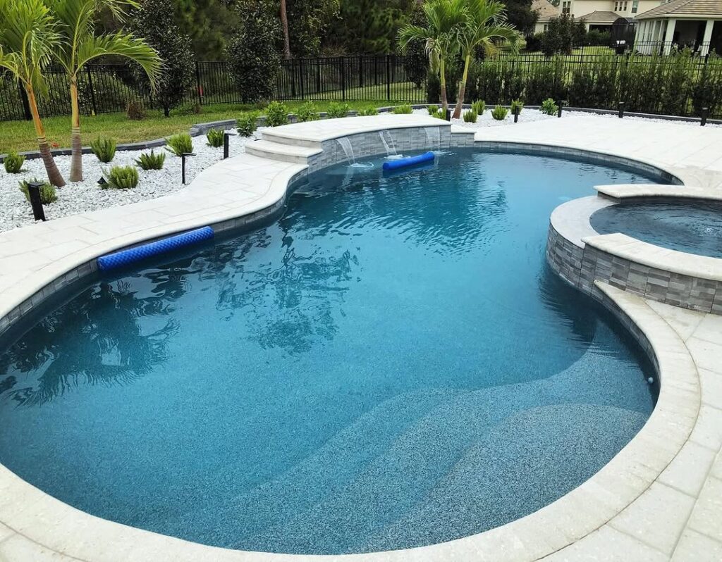 A backyard swimming pool with light gray stone decking, built-in steps, a raised hot tub, and surrounding landscaping with palm trees and shrubs by expert Pool Builders in Clermont.