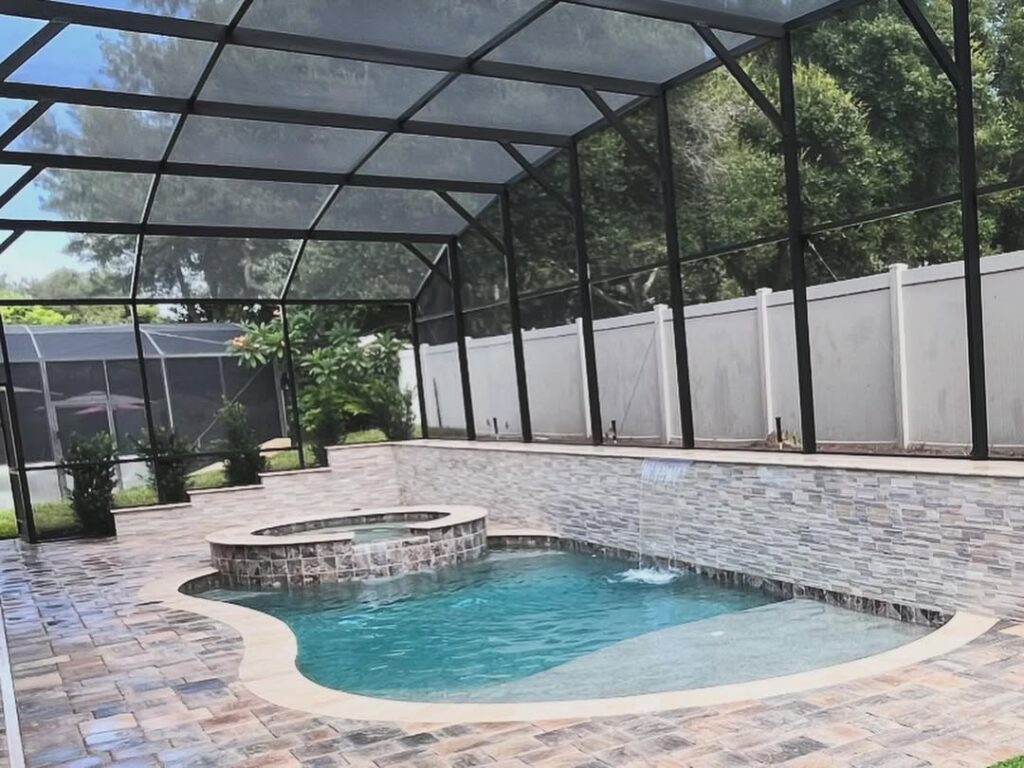 A small backyard pool with a connected hot tub, crafted by Pool Builders in Lake Nona, is surrounded by stone tiles and enclosed by a screened structure and a white fence.