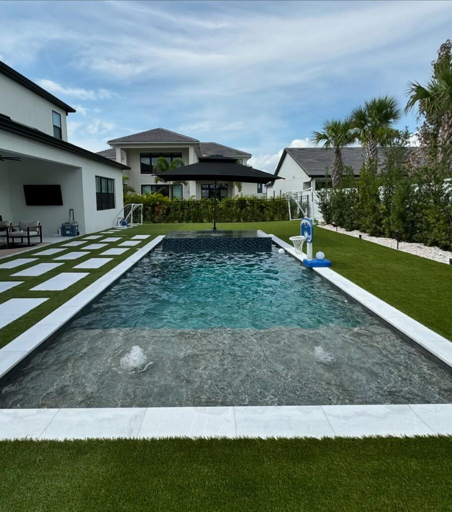 Rectangular backyard pool with a shallow entry, expertly crafted by Pool Builders in Clermont, surrounded by artificial grass and modern homes under a partly cloudy sky.