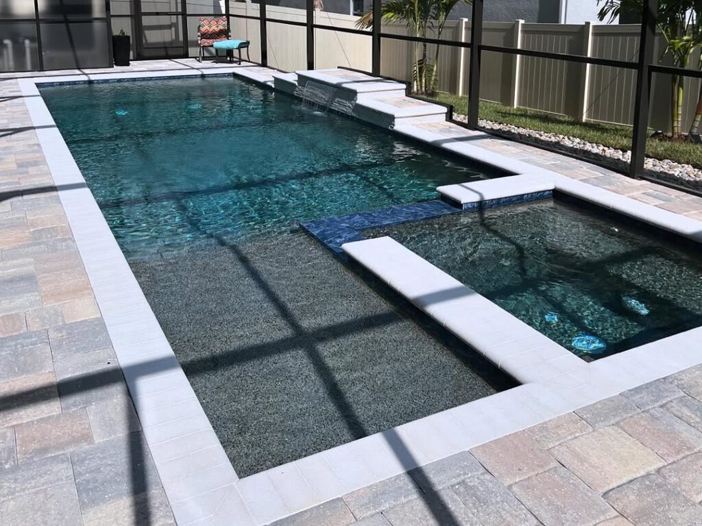 Rectangular backyard swimming pool with attached hot tub, crafted by expert Pool Builders in Clermont, surrounded by stone tile decking and enclosed by a screened fence.