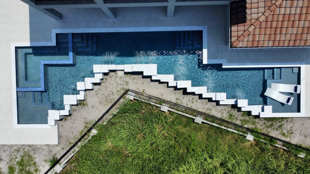 Aerial view of a modern, zigzag-shaped swimming pool next to a house with white decking, grass, and two poolside loungers.