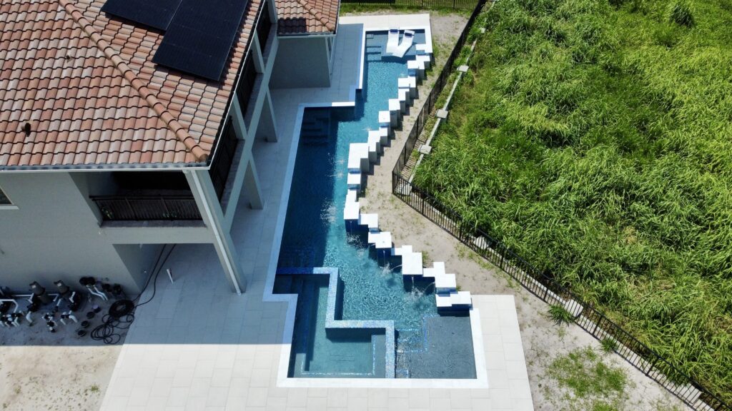 Aerial view of a modern backyard pool with a zigzag design and lounge chairs in the water, next to a tiled-roof house and a grassy area enclosed by a fence.