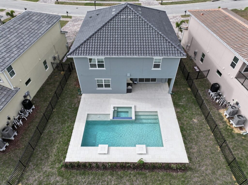 Aerial view of a two-story house with a fenced backyard featuring a rectangular swimming pool and attached spa, surrounded by neighboring homes.