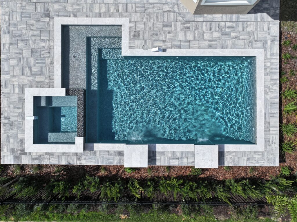 A rectangular backyard pool with an attached square hot tub, surrounded by gray stone tiles, viewed from above. Shrubs line the edge of the property.