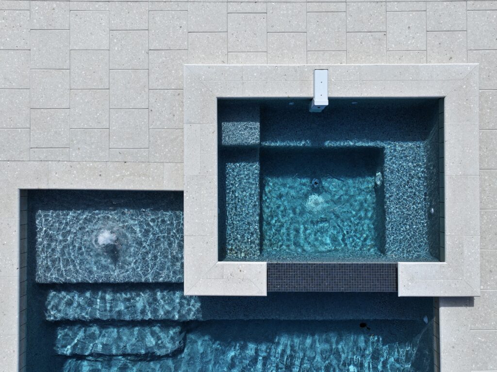 Aerial view of a rectangular hot tub with a waterfall spillway flowing into a swimming pool, both surrounded by light-colored stone tiles.