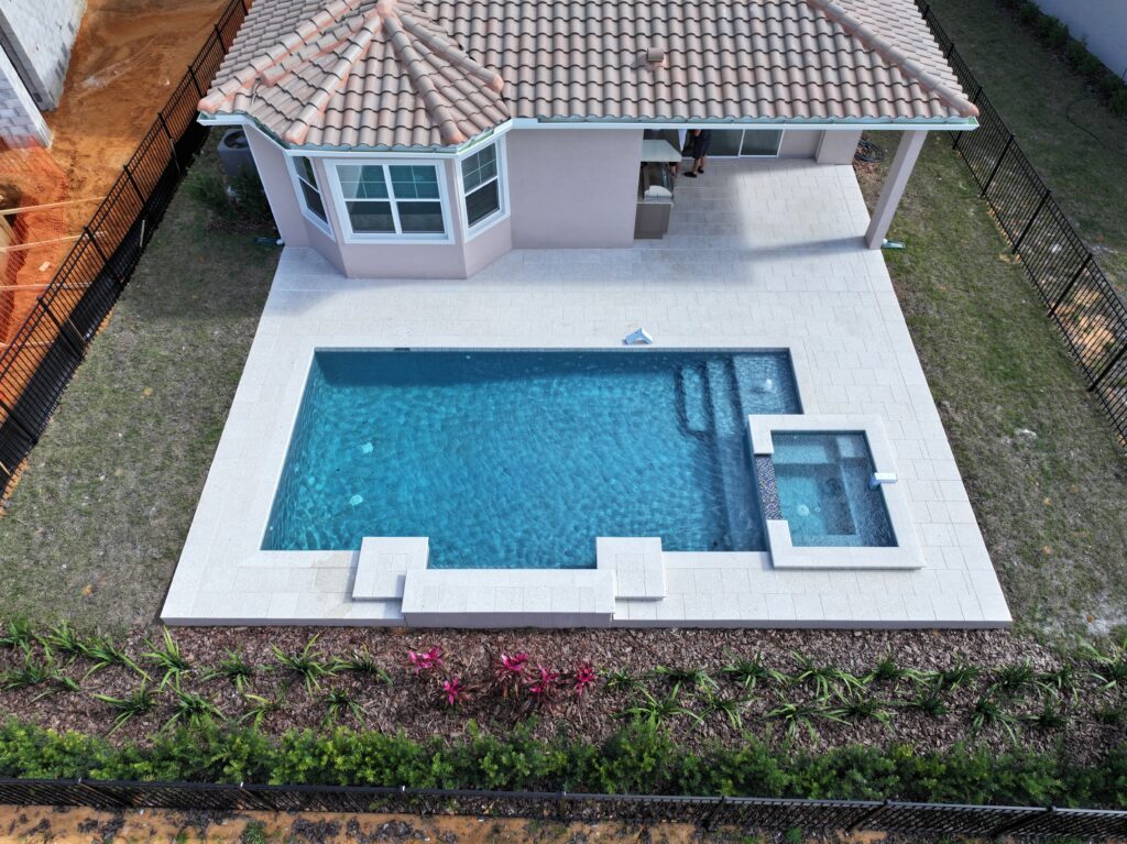 Aerial view of a rectangular backyard pool with attached hot tub, surrounded by a paved patio next to a house with a tiled roof and fenced yard.