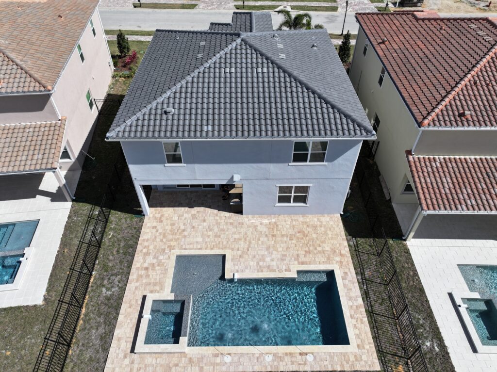 Aerial view of a two-story house with a tiled roof, fenced backyard, rectangular swimming pool, and attached hot tub, situated between two similar houses.