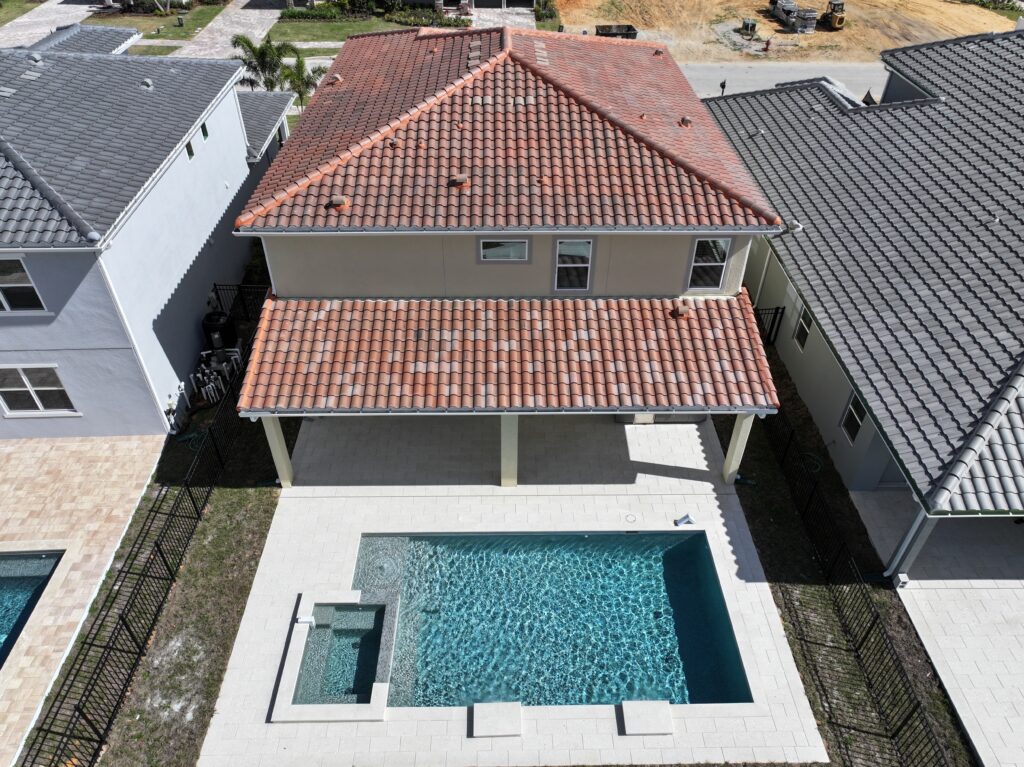 Aerial view of a two-story house with a covered patio and a rectangular backyard swimming pool with an attached spa, surrounded by a paved area and black metal fencing.