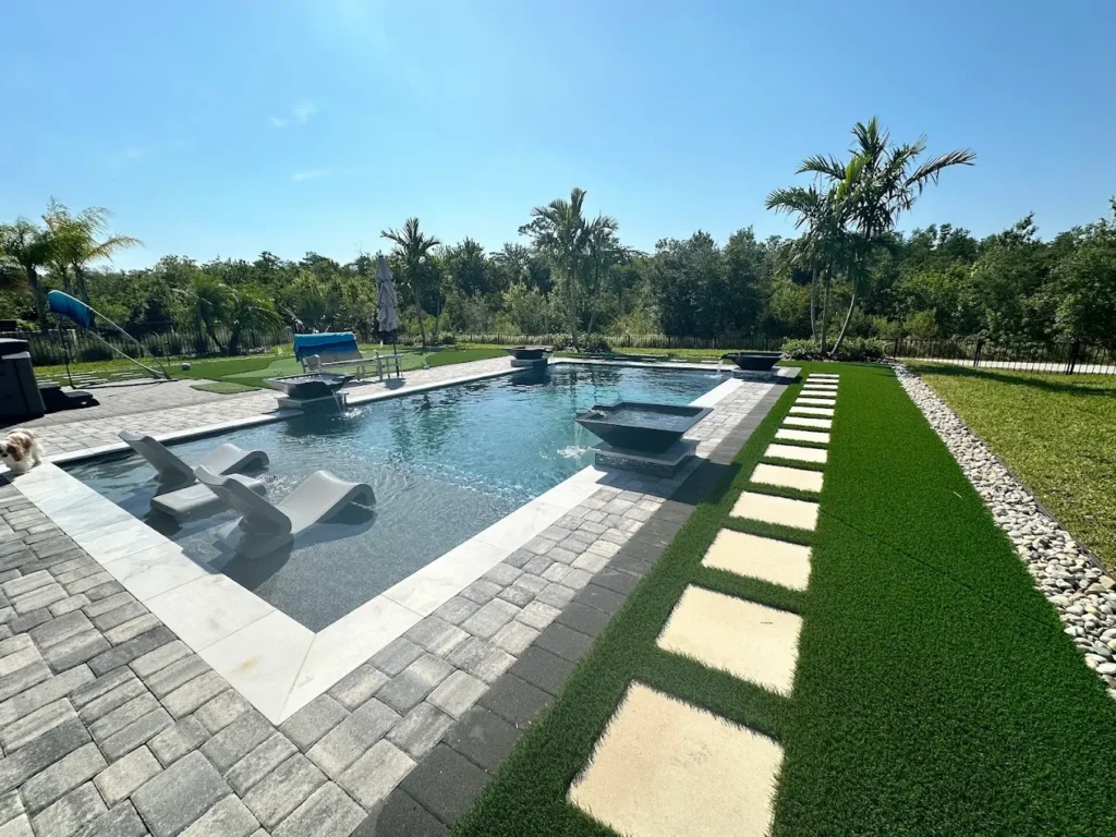 A rectangular swimming pool with in-pool lounge chairs, surrounded by stone pavers, artificial grass, stepping stones, and palm trees under a clear blue sky.