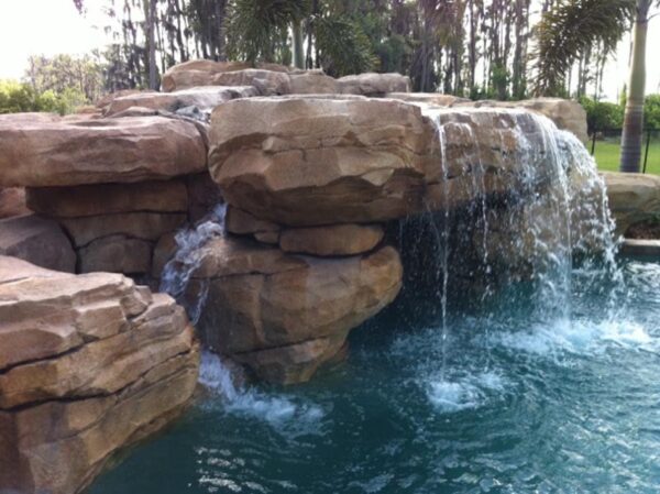 Large artificial rock waterfall flows into a clear swimming pool, surrounded by palm trees and greenery in the background.