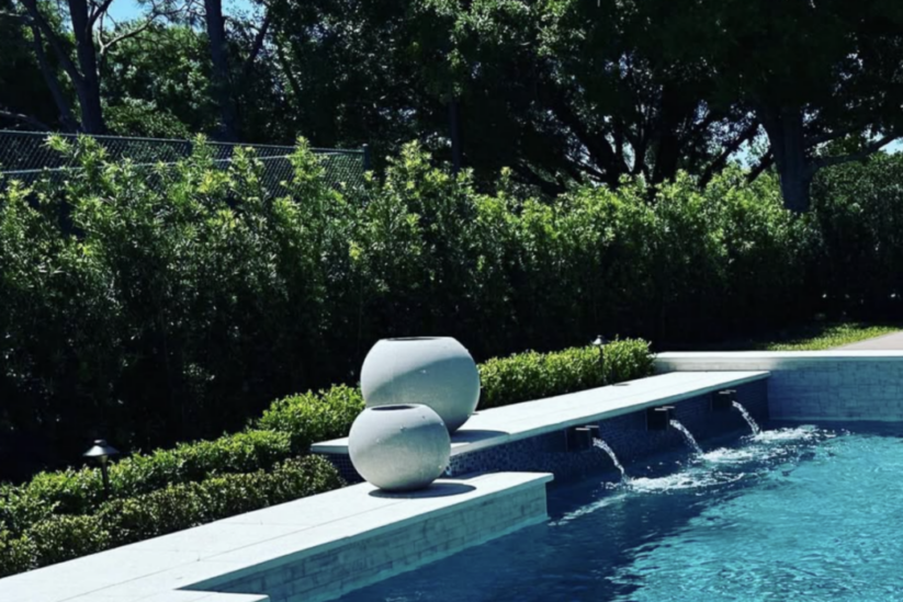 A modern pool with a bubbling hot tub in the foreground, three small waterfalls, and decorative white vases along the pool edge surrounded by greenery.