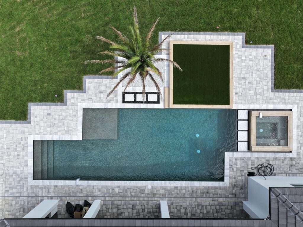 Aerial view of a rectangular swimming pool with adjacent hot tub, artificial turf area, a palm tree, and surrounding stone patio.