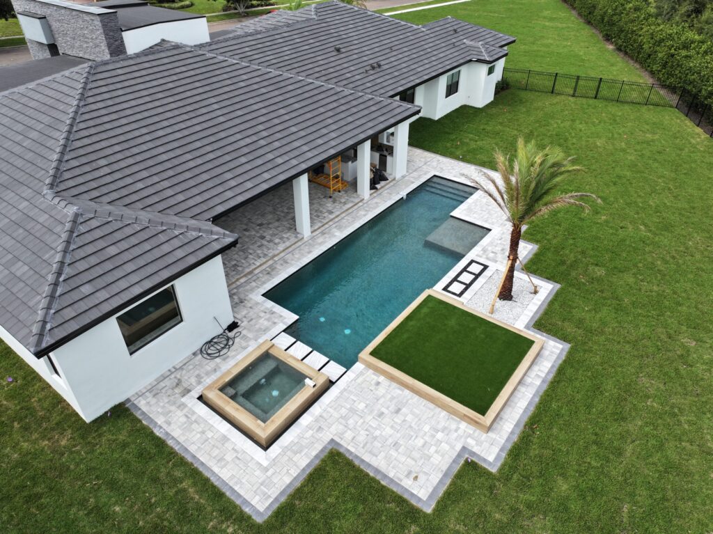 A modern house with a covered patio, rectangular swimming pool, hot tub, artificial turf area, and palm tree set in a grassy backyard.