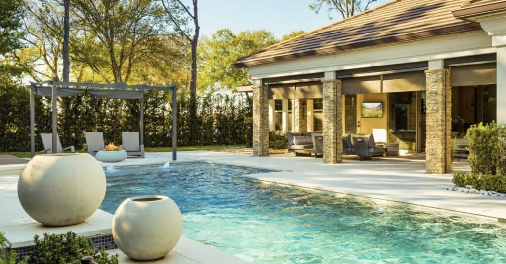 A backyard with a swimming pool, large white planters, lounge chairs under a pergola, and a covered patio with seating near a house.
