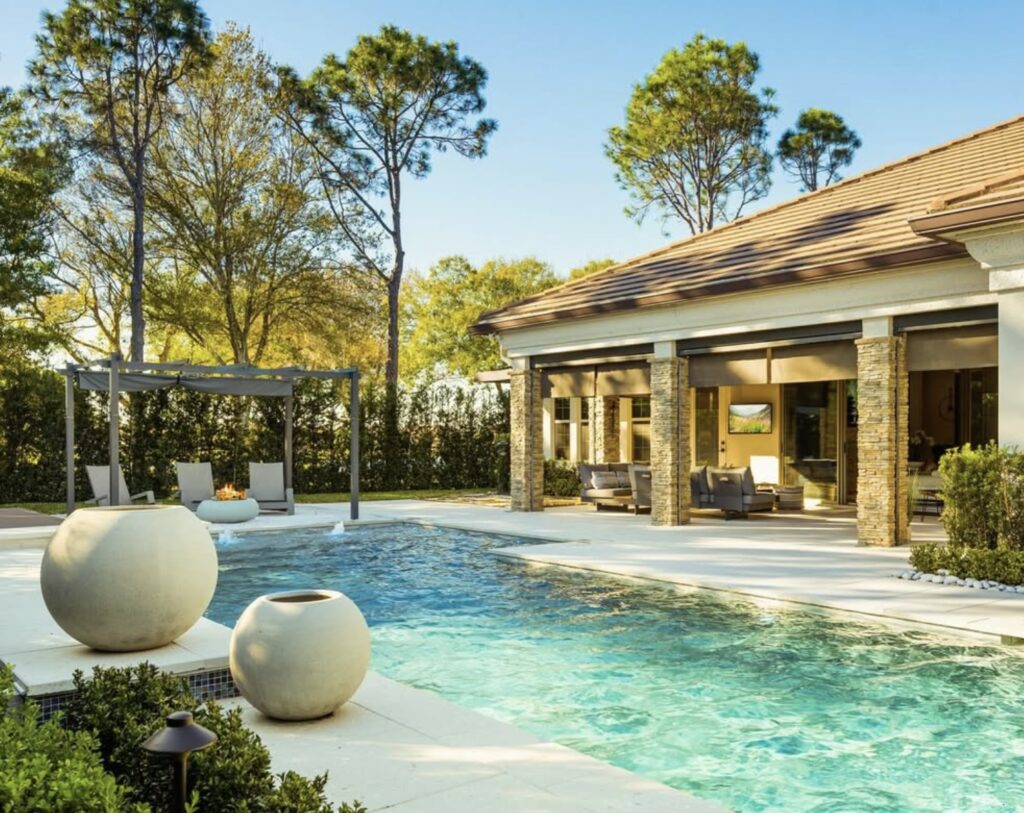 Modern backyard with a rectangular swimming pool, poolside lounge chairs under a pergola, decorative round planters, and a contemporary house with large windows and columns.