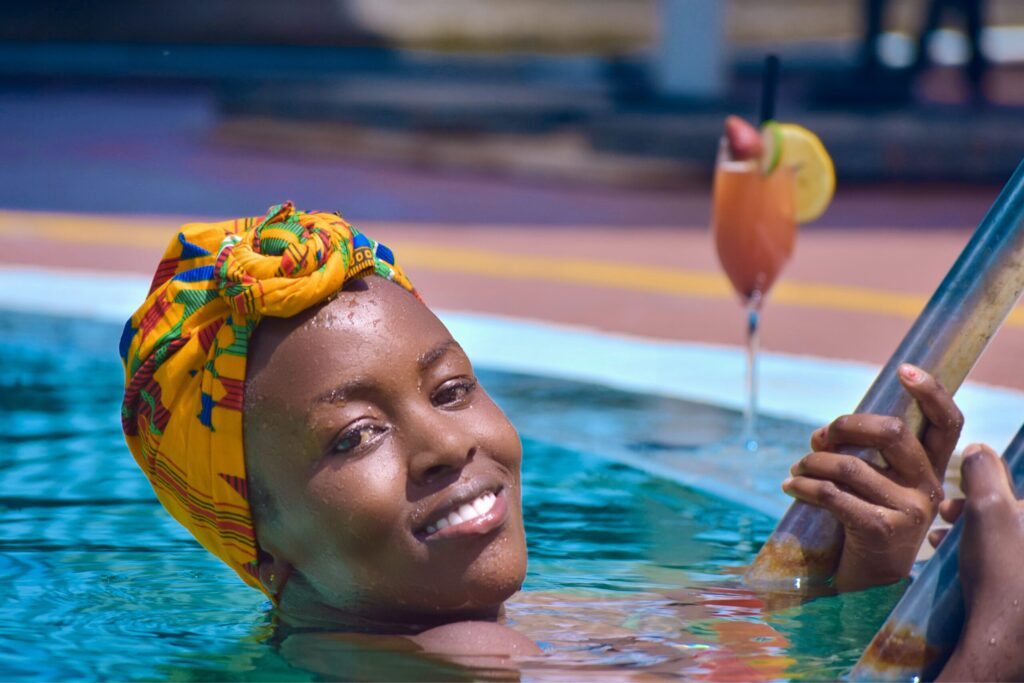 A person with a colorful headwrap smiles while holding a pool ladder in a swimming pool; a cocktail glass with garnish is on the poolside in the background.