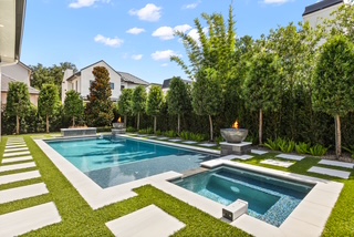 Modern backyard with a rectangular swimming pool, hot tub, and fire bowls, surrounded by green grass, trees, and large stepping stones on a sunny day.